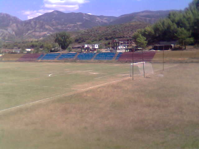 Empty soccer field with a goal, blue stadium bleachers, surrounding trees and mountains in the background, under a partly cloudy sky
