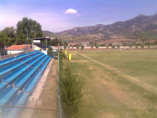 Soccer field viewed from behind blue stadium seating, with mountains, scattered buildings, and a cloudy sky in the distance